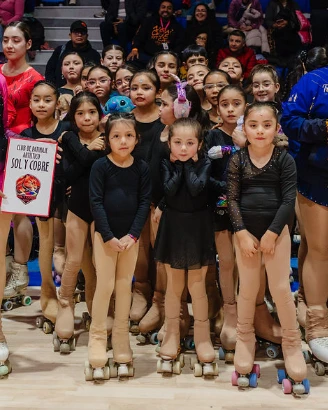 Grupo de deportistas del club con trajes de patinaje negros y patines, de pie junto a un cartel del club, con público de fondo en un gimnasio.