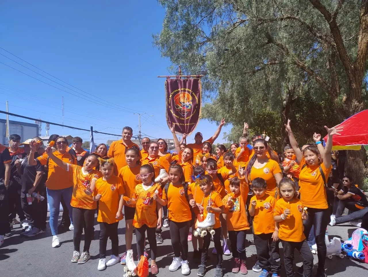 Niñas, niños, familias y equipo del club con poleras naranjas posando bajo el estandarte de Sol y Cobre en una calle, en un día soleado.